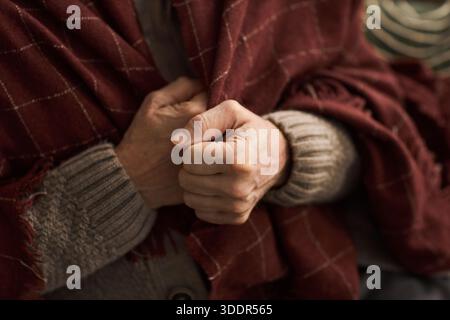 Senior Caucasian woman or man clasping hands together while sitting wrapped in blanket, close up of aged hands and textured fabric, emphasizing elderly female gesture during cold winter season Stock Photo