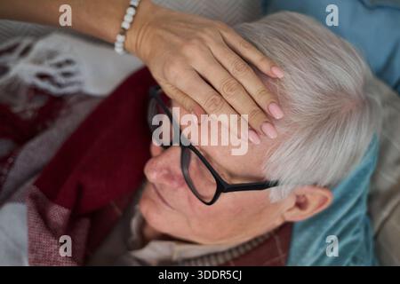 Senior Caucasian man lying in bed being cared for by middle aged Caucasian woman placing hand on his forehead checking temperature, close up of faces and hand Stock Photo