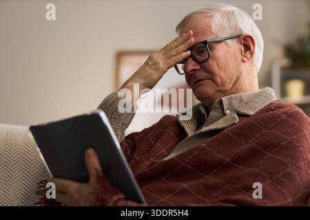 Senior Caucasian man wearing glasses sitting indoors holding tablet with one hand while touching forehead with other hand appearing unwell, concerned or thoughtful. suffering from cold or flu Stock Photo