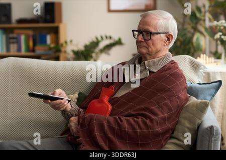Senior Caucasian man sitting on sofa holding remote control and hot water bottle, wearing glasses and blanket, watching television with focused expression in living room Stock Photo