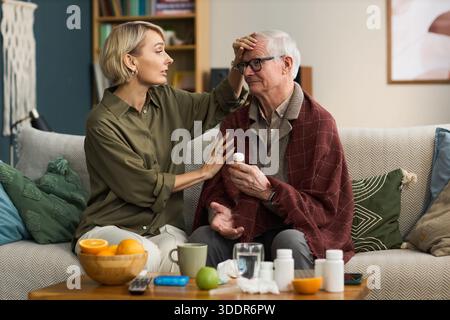Middle aged Caucasian woman checking temperature of senior Caucasian man sitting on sofa holding medicine bottle, various medication containers and fruit on table in foreground Stock Photo