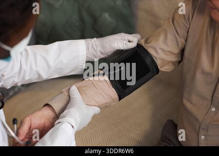 Middle aged Black man wearing medical coat measuring blood pressure of senior Caucasian man using digital sphygmomanometer, hands and partial faces visible, clinical setting Stock Photo