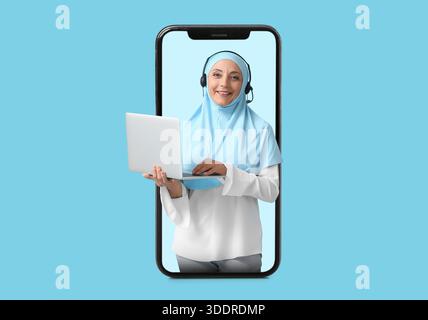 Female Muslim technical support agent with laptop popping out of smartphone screen on blue background Stock Photo