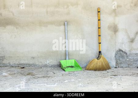 A traditional grass broom with a golden handle and a modern green plastic dustpan, holding debris, lean against a textured, light grey concrete wall a Stock Photo