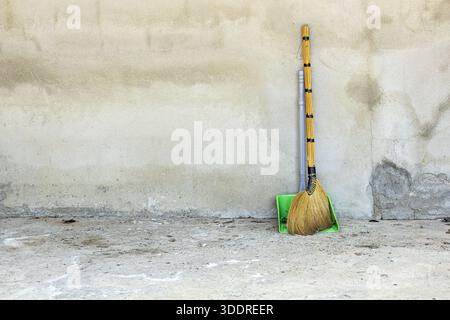 A traditional grass broom with a black-wrapped handle and a bright green plastic dustpan lean against a textured, light gray concrete wall. The dusty, Stock Photo