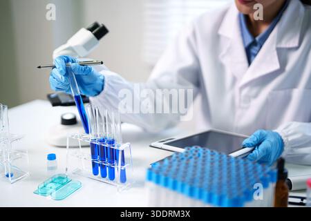 Adult researcher wearing protective gloves examines blue liquid in test tubes during a scientific laboratory experiment using a digital tablet. Stock Photo
