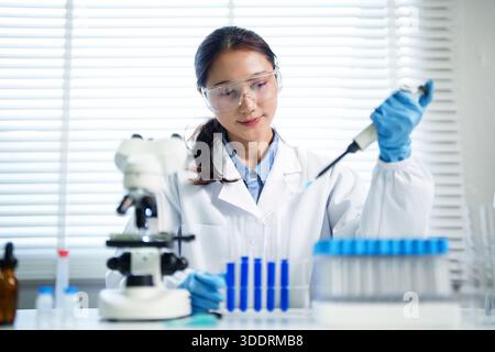 Professional Asian woman scientist performs precise lab experiment wearing protective goggles and gloves handling chemical samples Focus on research Stock Photo