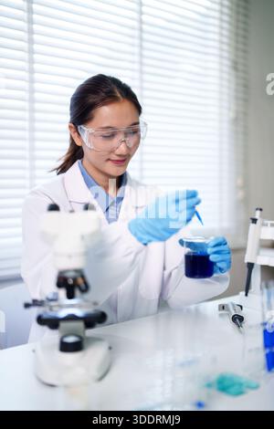 Adult Asian woman performs chemistry experiment in laboratory wearing safety goggles gloves holding beaker pipette demonstrating scientific method Stock Photo