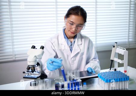 Focused Asian adult woman scientist performs lab experiment with blue liquid chemicals using a tablet and microscope in modern research facility Stock Photo