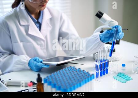 Asian adult scientist performs crucial lab experiment with test tubes and tablet. Precision work in modern research facility. Stock Photo