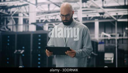 IT Professional Inspecting Racks in Data Center. Bearded Male Inspecting or Troubleshooting Equipment With Handheld Device Amidst Rows Data Servers. C Stock Photo