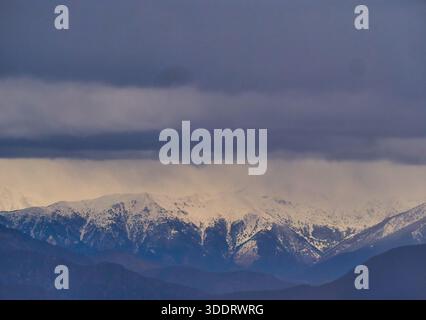 Snow-Covered Mountain Range Under Heavy Dark Clouds at Dusk, Serene Winter Landscape Stock Photo
