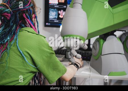 Woman and humanoid robot arm with articulated fingers above computer desk in modern tech workspace, working together, pregaming and operating  Stock Photo