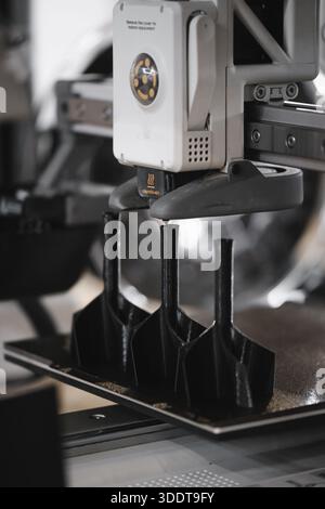 Industrial 3D printer extruder head printing multiple black plastic parts on build plate during additive manufacturing process. Close-up of heated 3D Stock Photo