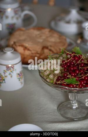 Glass bowl with red and white currants stands on a tea table with homemade apple charlotte and floral teaware softly blurred in the background Stock Photo