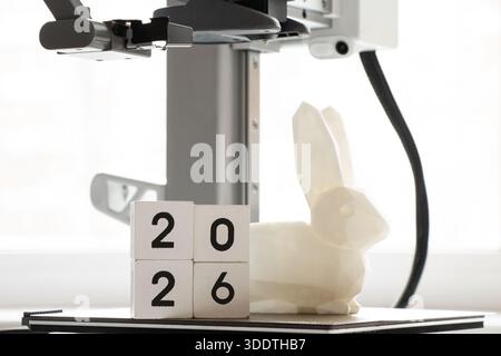 A desktop 3D printer and a white low-poly bunny on the printer, female hands, and cubes with the year 2026, modern technologies for additive manufactu Stock Photo