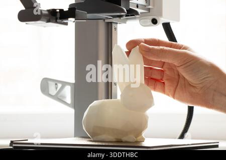 A desktop 3D printer and a white low-poly bunny on the printer, along with female hands, modern technologies for additive manufacturing Stock Photo