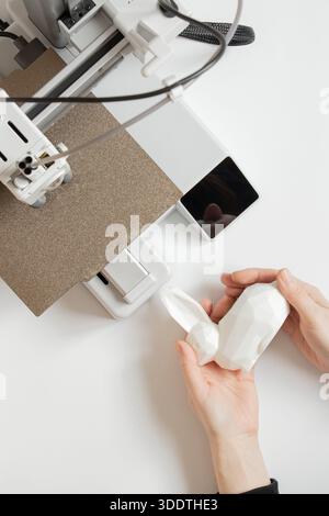 A desktop 3D printer and a white low-poly bunny on the printer, along with female hands, modern technologies for additive manufacturing Stock Photo