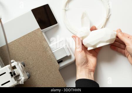 A desktop 3D printer and a white low-poly bunny on the printer, along with female hands, modern technologies for additive manufacturing Stock Photo