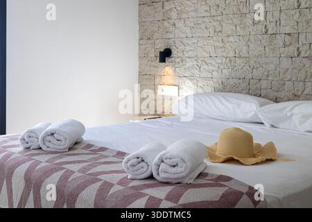 Inviting bedroom setup with textured wall, white linens, towels and straw hat on bed Stock Photo