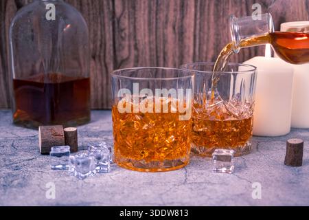 Whisky is poured into a second crystal glass while another drink rests on the table, with bottle and candlelight. Bar service scene for celebration, t Stock Photo