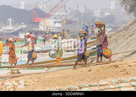 Dhaka, Bangladesh - 19 February 2025: View of laborers walking on makeshift planks near the Buriganga River, carrying heavy loads under a hazy sky, wi Stock Photo
