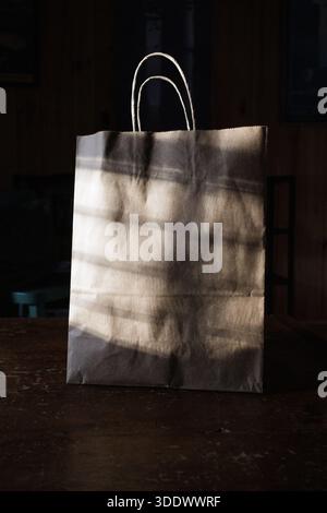 Dappled sunlight creates soft shadows on a paper shopping bag with handles, highlighting its minimalist and eco-friendly design. Stock Photo