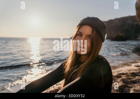 Woman, beanie, sunset, young female sitting on a rocky beach enjoying the golden hour ocean tranquility. Stock Photo