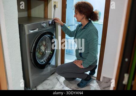 Woman kneels to select program settings on modern washing machine control panel at home Stock Photo