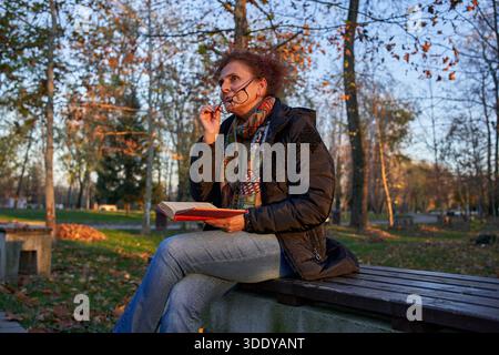 Woman removes glasses and looks into the distance while holding book on autumn park bench Stock Photo