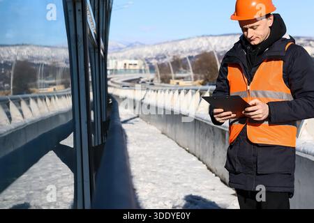 Side view of construction worker in orange helmet and vest operating tablet on icy highway bridge with mountain view. Stock Photo