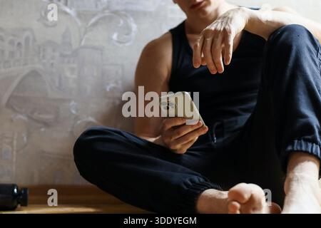 Muscular barefoot man in dark casual clothes rests on wooden floor against wall, holding and looking at smartphone screen with contemplative expressio Stock Photo