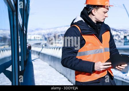 Profile view of helmeted man in orange vest holding and reading clipboard on a winter bridge with mountainous backdrop. Stock Photo