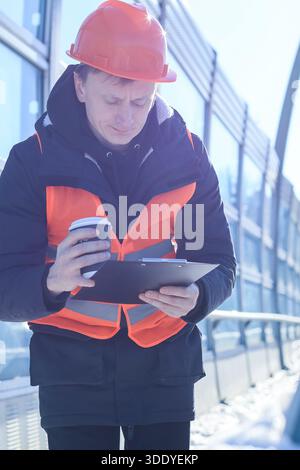 Male worker in orange hardhat holds disposable coffee cup and examines notes on clipboard in winter outdoor setting. Stock Photo
