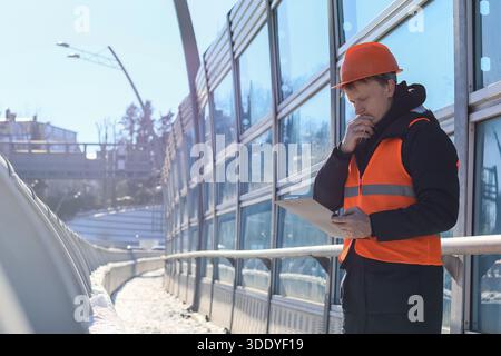 A contemplative man in protective clothing holds clipboard while thinking beside snowy noise screen on urban bridge structure. Stock Photo