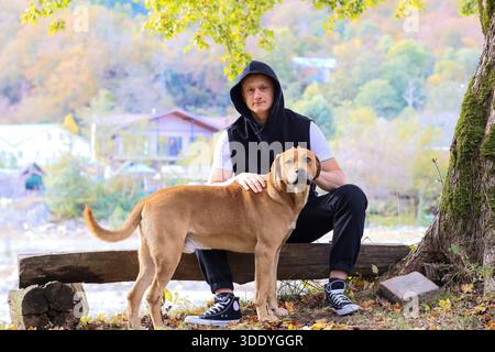 A young man in a black hoodie and pants sits cross-legged on a wooden log by the river, hands clasped in contemplation, amidst golden autumn foliage a Stock Photo