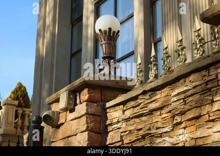 Elegant cast bronze outdoor lamp with diffused white globe atop rough stone plinth, surveillance camera and cypress tree in background. Stock Photo