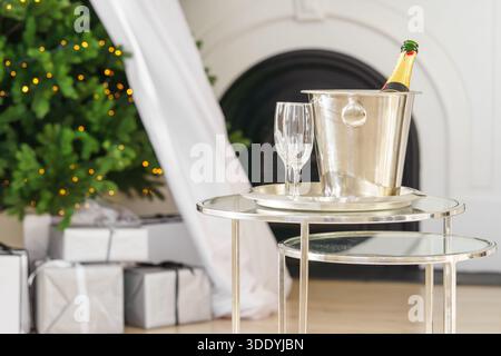 Champagne bottle in metal ice bucket with two empty glasses on glass table on glowing Christmas tree lights and gifts, present background. Festive win Stock Photo