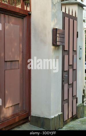 Rustic red metal pedestrian gate featuring abstract rectangular panels, mounted intercom, and spiked top on a stucco wall. Stock Photo