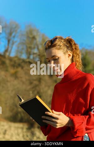 A young woman with ponytail and sunglasses on head holds an open book while standing in a natural landscape with trees and clear sky. Stock Photo