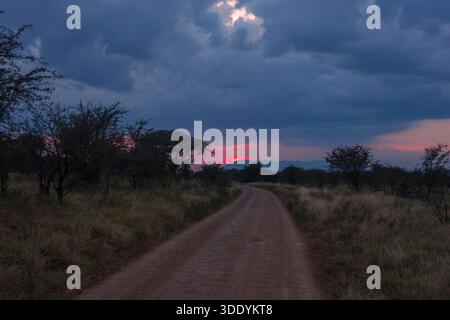 The sun sets behind the mountains with a dirt road leading up to the horizon with moody, dark clouds at Central Serengeti National Park, Tanzania. Stock Photo