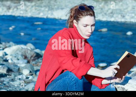 Young woman sits on stones by blue river, engrossed in open book with sunglasses on head. Stock Photo