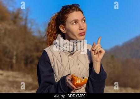 Laughing young woman peeling mandarin, joyful moment in sunny autumn mountain forest Stock Photo