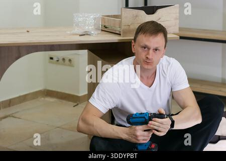 A focused man in casual clothes sits on the floor using a blue cordless drill to assemble light wooden shelves and drawers in a bright room. Stock Photo