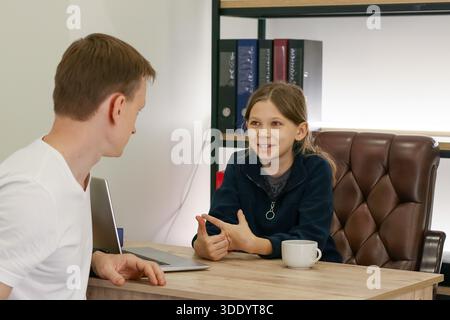 Adult male talks while child gestures expressively at office table with laptop, cup, and background shelves Stock Photo