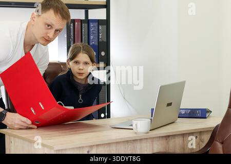 Adult male extends red binder across table to attentive child in professional space with shelves and laptop Stock Photo