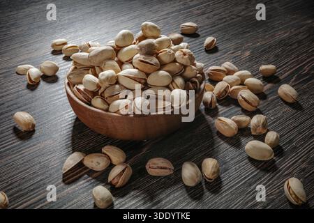 Wooden bowl filled with pistachios on dark wooden table, tasty salted nuts scattered around, healthy snack and natural food concept. Stock Photo
