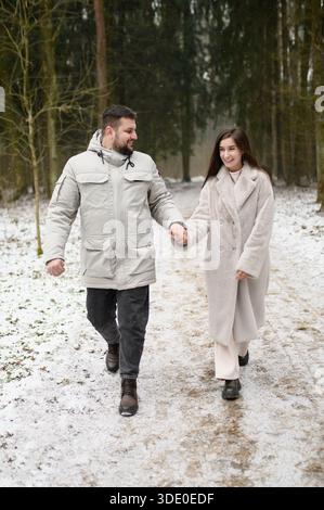 Loving couple enjoys winter vacation, walking through snowy forest, holding hands. Man holds beloved's hand, enjoying walk. Winter vacation in snowy forest. Valentine's Day. Christmas morning Stock Photo