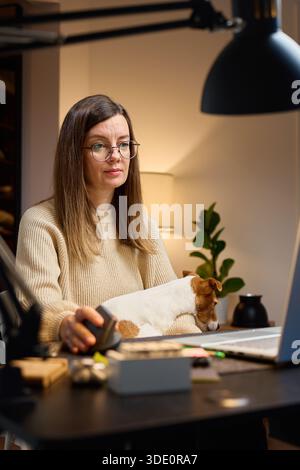 Woman sitting at home desk, working on laptop at home office with small dog on her lap. Person using computer at room with jack russell terrier. Life with pet concept Stock Photo