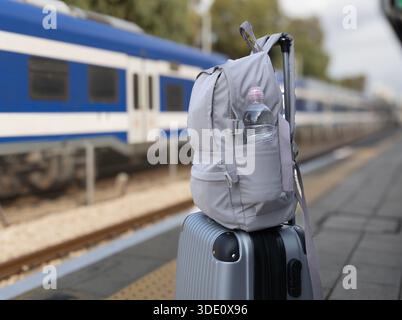 Horizontal wide angle eye level shot of a silver travel luggage and a grey backpack standing on a railway station as a blue train passes by Stock Photo